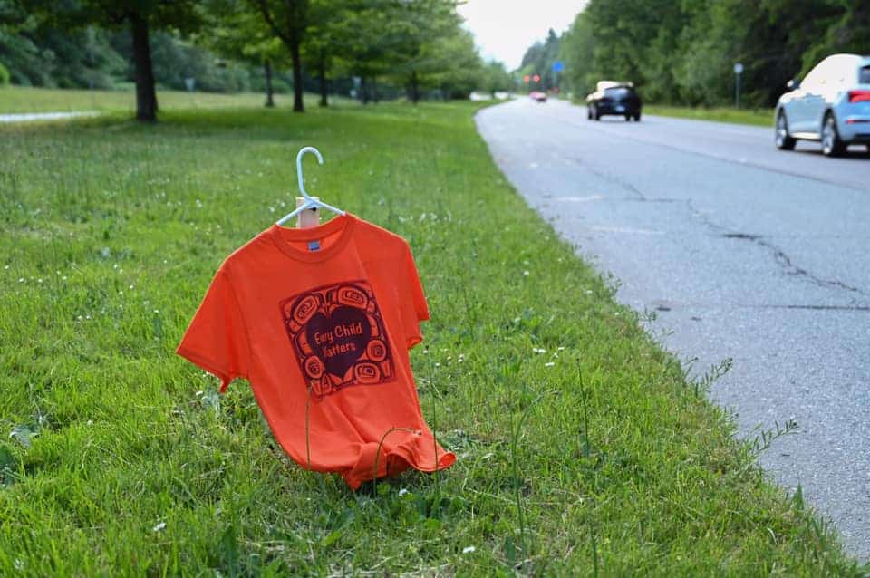 An orange shirt that says "Every Child Matters" hangs on a white clothes hanger which is held up by a wooden stake. It is placed on a grassy boulevard, with cars driving on the right hand side.